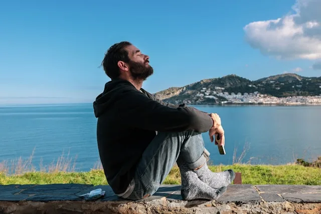 A man by the sea with his phone lowered, eyes closed — letting the screen go and reaching for something real