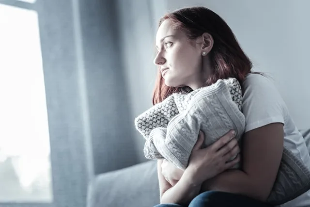 A young woman sitting alone by a window holding a cushion — young adults report the highest loneliness of any generation