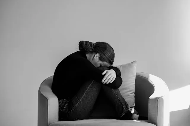 A person curled up on a chair with their head resting on their knees, looking withdrawn and alone