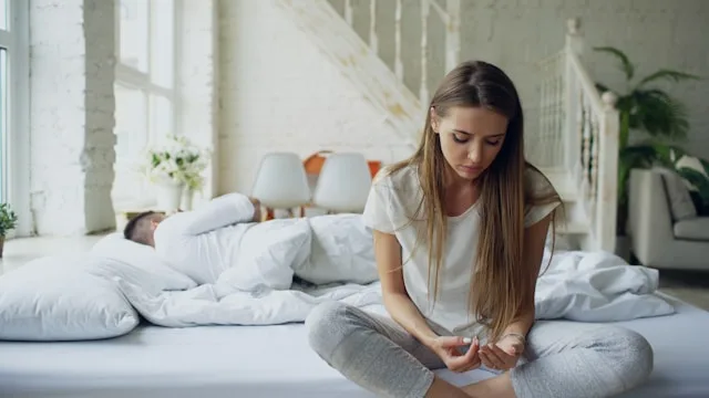 A woman sitting on the edge of the bed looking down while her partner lies turned away behind her, feeling distant