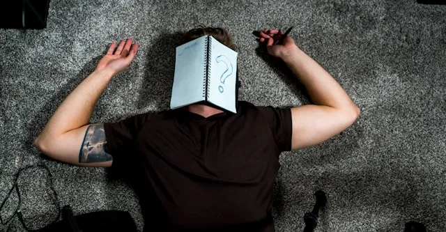 A person lying down with an open notebook showing a question mark over their face — asking why they feel so lonely